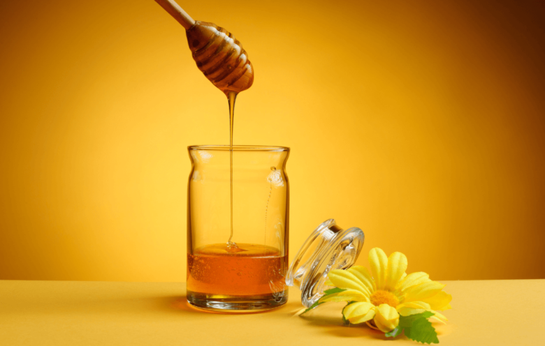 imagem com fundo amarelo, no centro um pote de mel e ao lado uma flor